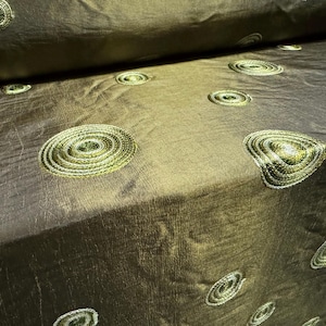 May include: A close-up of a brown fabric with a subtle sheen. The fabric is decorated with a repeating pattern of gold embroidered circles.