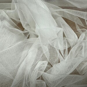 May include: White tulle fabric with a fine mesh texture. The fabric is draped and folded, creating a soft and delicate look.