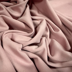 Può includere: Primo piano di un tessuto rosa cipria con morbide pieghe e una superficie strutturata. Il tessuto è drappeggiato, creando ombre e luci che ne enfatizzano la natura fluida. Il materiale sembra essere un tessuto di peso medio, adatto all'abbigliamento o all'arredamento.