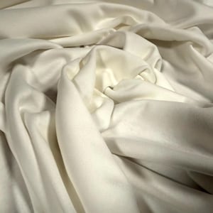 May include: Close-up of off-white fabric with soft folds and shadows. The fabric appears to be a lightweight material, possibly a knit or woven textile, with a smooth texture. The image highlights the drape and flow of the fabric.
