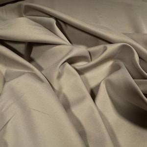 Op de afbeelding: Een close-up van een gladde, beige stof met een zachte, soepele textuur. De stof is gevouwen en zo gerangschikt dat de luxe uitstraling wordt benadrukt.