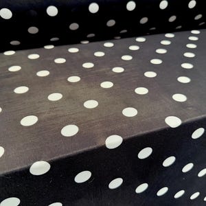 Può includere: Una lunghezza di tessuto nero piegato con un motivo ripetitivo di pois bianchi. Il materiale ha una consistenza liscia e leggermente opaca ed è disposto a strati, mostrando le dimensioni e la spaziatura costanti della stampa circolare.