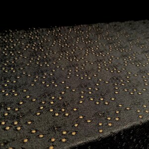 May include: A rectangular black fabric surface featuring a dense, repeating pattern of small, circular gold-colored perforations. The material has a textured, slightly matte finish, and the gold dots are arranged in a scattered, organic layout.