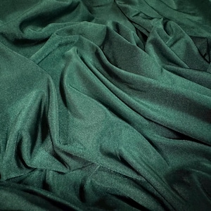 May include: A close-up shot of a dark emerald green fabric with soft folds and a subtle texture. The material appears to be a knit or woven textile, with a slight sheen. The fabric is draped, creating shadows and highlights.