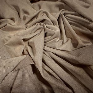 Könnte beinhalten: Ein Stück beigefarbener Netzstoff mit einem feinen, gleichmäßigen Gittermuster. Das Material ist in weichen, fließenden Falten drapiert, wodurch eine strukturierte Oberfläche mit wechselndem Licht und Schatten entsteht, die die Transparenz betont.