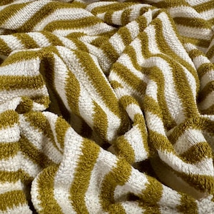 Op de afbeelding: Close-up van een gevouwen, getextureerde handdoek met afwisselende strepen van olijfgroen en wit. De stof ziet er zacht en pluche uit, met een licht gekreukte uitstraling. De handdoek is waarschijnlijk gemaakt van katoen of een vergelijkbaar absorberend materiaal.