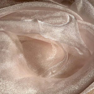 May include: Close-up of a shimmering, light pink fabric with a soft, flowing texture. The material appears to be organza or a similar sheer textile, with subtle highlights and shadows creating depth. The fabric is gathered and draped.