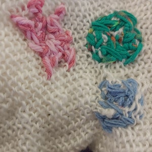 Op de afbeelding: Wit gebreid stof met drie geborduurde plekken: een roze ster, een groene cirkel en een blauwe bloem.