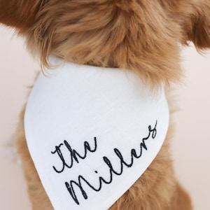 May include: A white dog bandana with the embroidered text "The Millers" in black script. The bandana is around a dog's neck, with the dog's brown fur visible.