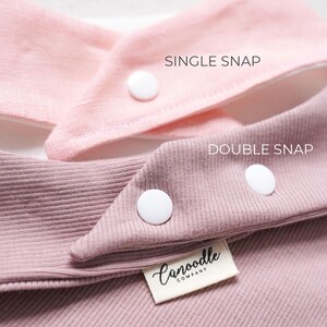 May include: Two ribbed fabric bibs in light pink and dusty rose. The light pink bib features a single white snap, labeled SINGLE SNAP. The dusty rose bib features two white snaps, labeled DOUBLE SNAP, and a white tag reading Canoodle COMPANY.