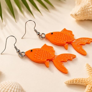 May include: A pair of bright orange goldfish earrings featuring a textured scale pattern, a small black eye, and a detailed fin design. The fish charms are attached to silver-toned metal fishhook ear wires, creating a dangling style for the wearer.