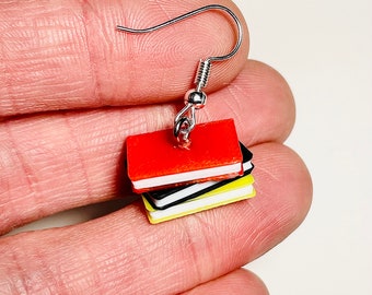 May include: A dangle earring featuring a miniature stack of three books in red, black, and yellow. The books have white pages visible along the edges and are attached to a silver-toned metal fishhook ear wire for pierced ears.
