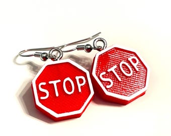 May include: A pair of dangle earrings featuring octagonal red stop signs with white borders and the word STOP in white lettering. The charms are attached to silver-toned metal fishhook ear wires, creating a distinct and recognizable traffic sign design.