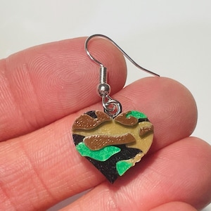 May include: Camouflage heart-shaped earrings with a silver hook. The heart is made of a green, brown, and black camouflage pattern.