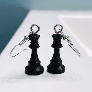 May include: A pair of black chess queen earrings featuring a textured, layered finish. Each piece is attached to a silver-toned metal hook with a small bead and coil detail, designed for hanging from the earlobe with a classic drop style.
