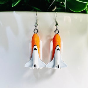 May include: A pair of silver earrings featuring a white and orange space shuttle design.