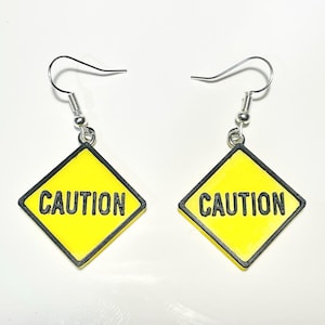 May include: A pair of dangle earrings featuring a diamond-shaped yellow charm with a black border and the word CAUTION printed in black capital letters. The charms are attached to silver-toned metal fishhook ear wires for pierced ears.