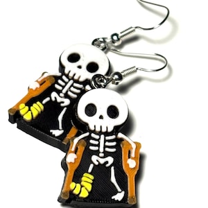 May include: A pair of Halloween-themed earrings featuring a skeleton design. The skeletons are white with black accents, wearing orange crutches and yellow casts. The earrings have silver-colored fish hook ear wires and a black background.