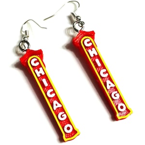 Op de afbeelding: Een paar hangende oorbellen met lange, rechthoekige rode hangers met gele randen. Het woord CHICAGO staat verticaal in witte hoofdletters op elke hanger gedrukt. De oorbellen zijn bevestigd aan zilverkleurige metalen oorhaakjes.