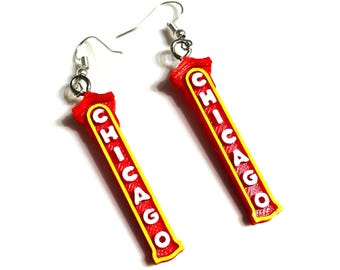 May include: A pair of dangle earrings featuring long, rectangular red pendants with yellow borders. The word CHICAGO is printed vertically in white capital letters on each pendant. The earrings are attached to silver-toned metal fishhook ear wires.