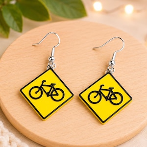 May include: A pair of dangle earrings featuring a diamond-shaped yellow charm with a black bicycle graphic, mimicking a road sign. The charms are attached to silver-toned metal fishhook ear wires, providing a secure and simple way to wear these items.
