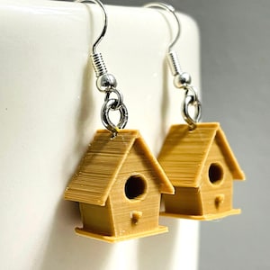 May include: A pair of dangle earrings featuring miniature light brown birdhouse charms. Each birdhouse has a triangular roof, a circular entrance hole, and a small perch. The charms are attached to silver-toned metal fishhook ear wires.