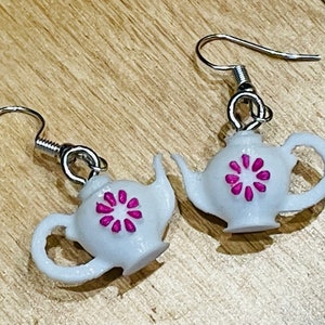 May include: A pair of white teapot earrings with pink flower accents. The earrings are made of polymer clay and have silver hooks.