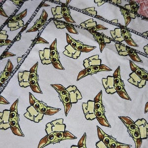 Puede incluir: Varios pa&ntilde;os blancos cuadrados peque&ntilde;os con un patr&oacute;n repetido de un personaje de dibujos animados. Los pa&ntilde;os tienen un borde cosido negro. Una botella amarilla es parcialmente visible en el fondo.