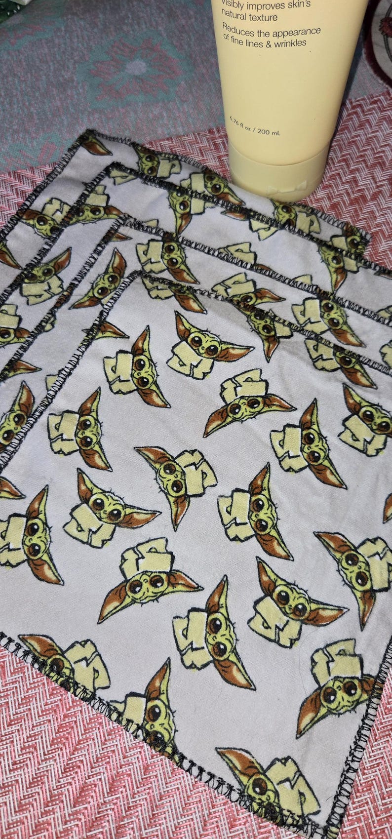 Puede incluir: Varios pa&ntilde;os blancos cuadrados peque&ntilde;os con un patr&oacute;n repetido de un personaje de dibujos animados. Los pa&ntilde;os tienen un borde cosido negro. Una botella amarilla es parcialmente visible en el fondo.