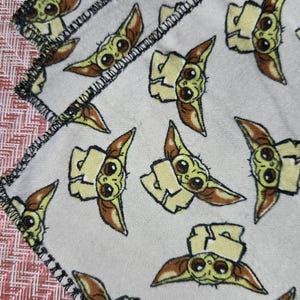Puede incluir: Manta blanca con un patr&oacute;n repetido de un personaje de dibujos animados con ojos grandes y orejas puntiagudas. El personaje es de color verde claro y marr&oacute;n. La manta tiene un borde cosido negro y un borde con estampado rojo y blanco.