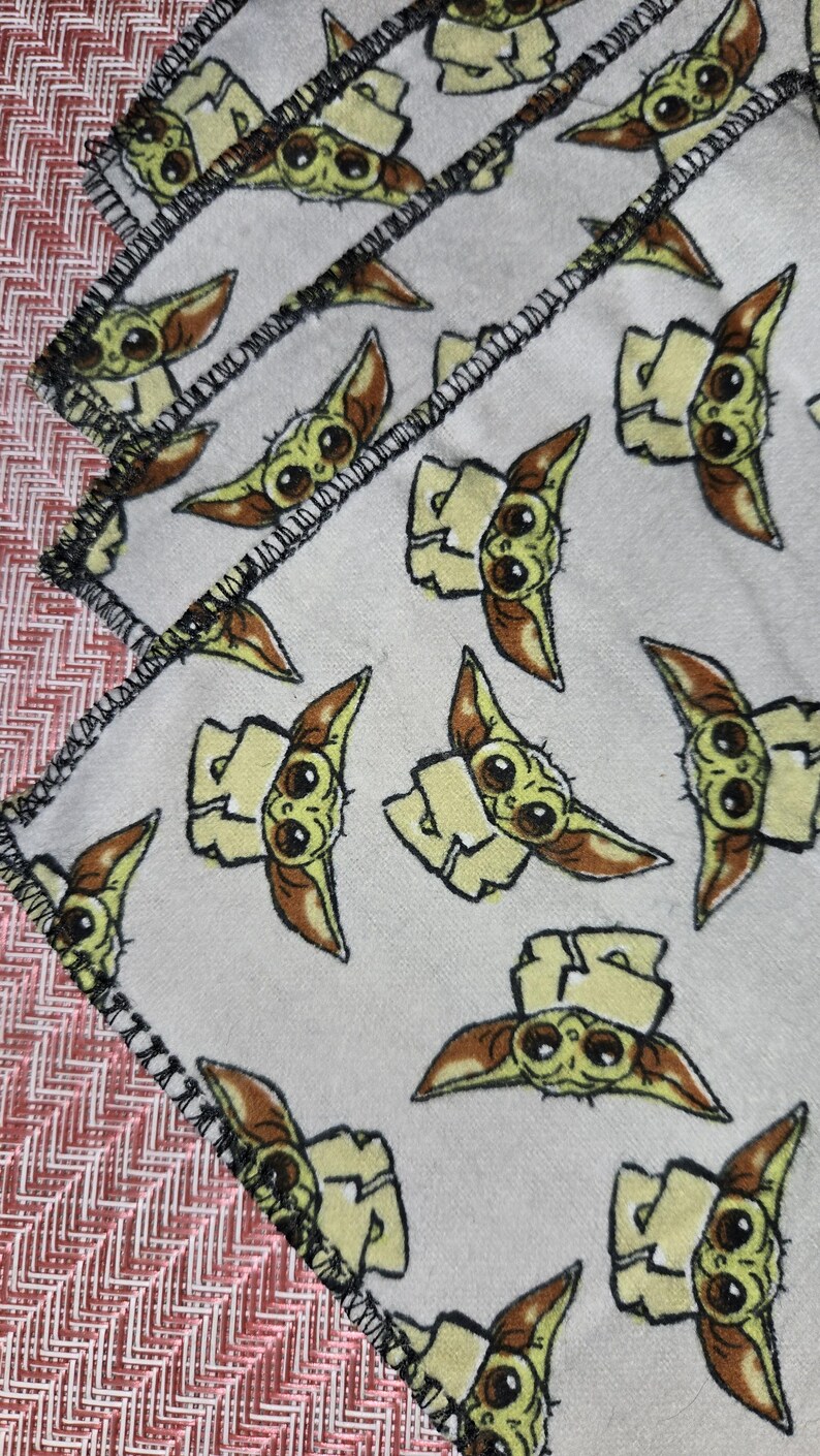 Puede incluir: Manta blanca con un patr&oacute;n repetido de un personaje de dibujos animados con ojos grandes y orejas puntiagudas. El personaje es de color verde claro y marr&oacute;n. La manta tiene un borde cosido negro y un borde con estampado rojo y blanco.