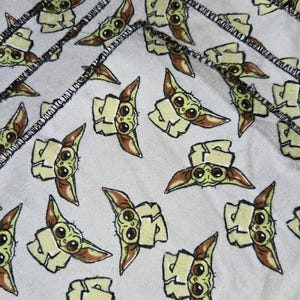 Puede incluir: Servilletas de tela blancas con un patr&oacute;n repetido de un personaje de dibujos animados con ojos grandes y orejas puntiagudas. El personaje es de color verde claro, marr&oacute;n claro y marr&oacute;n. Los bordes de las servilletas est&aacute;n cosidos con hilo negro.