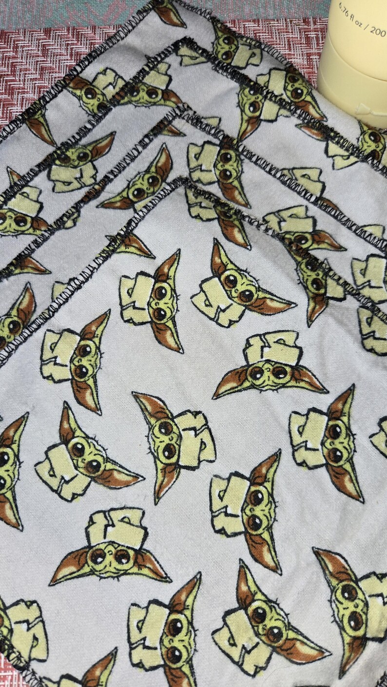 Puede incluir: Servilletas de tela blancas con un patr&oacute;n repetido de un personaje de dibujos animados con ojos grandes y orejas puntiagudas. El personaje es de color verde claro, marr&oacute;n claro y marr&oacute;n. Los bordes de las servilletas est&aacute;n cosidos con hilo negro.