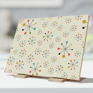 May include: A white ceramic tile with a repeating pattern of colorful mid-century modern starburst designs. The tile is on a wooden stand.