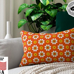 May include: Orange throw pillow with a white daisy pattern. The pillow is on a couch with a white blanket. The text "Motor City Design Works" is on a black and white sticker in the upper right corner of the image.