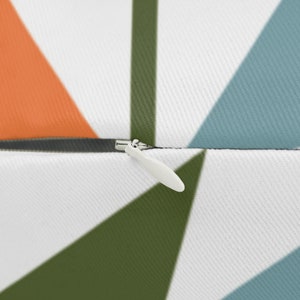 Op de afbeelding: Close-up van een wit kussen met een geometrisch patroon. Het ontwerp heeft oranje, blauwe en groene driehoeken tegen een witte achtergrond. Een witte rits is zichtbaar aan de zijkant.