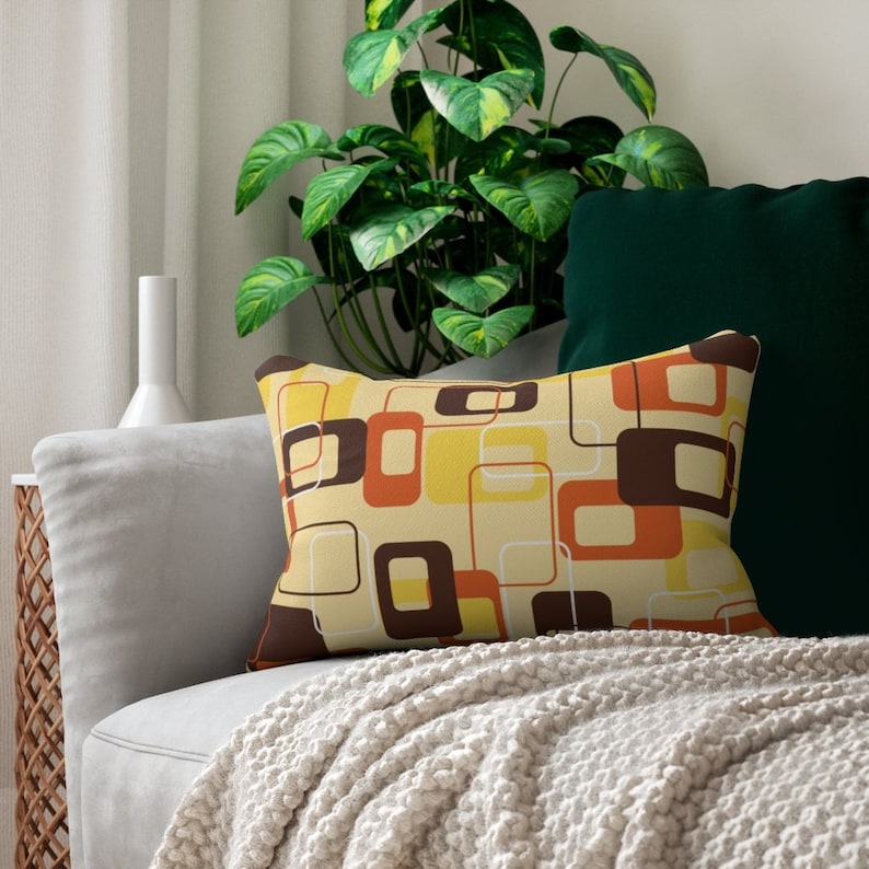 K&ouml;nnte beinhalten: Ein Dekokissen mit einem geometrischen Muster aus braunen, orangen und gelben Quadraten auf cremefarbenem Hintergrund. Das Kissen liegt auf einem hellgrauen Sofa mit einer wei&szlig;en Wolldecke.