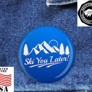 Op de afbeelding: Een blauwe knoop met witte tekst en afbeeldingen. De knoop heeft een bergketen, bomen en de tekst "Ski You Later!". De knoop heeft ook de tekst "Printed in the USA" en een logo.