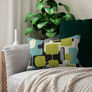 May include: A decorative cushion with a geometric pattern in shades of green, blue, black, and white. The cushion is on a light grey sofa with a white throw blanket.