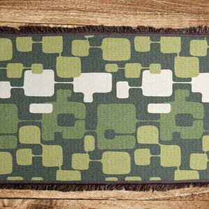 May include: A woven throw blanket with a geometric pattern of green, white, and gray squares and rectangles. The blanket has a fringe border.