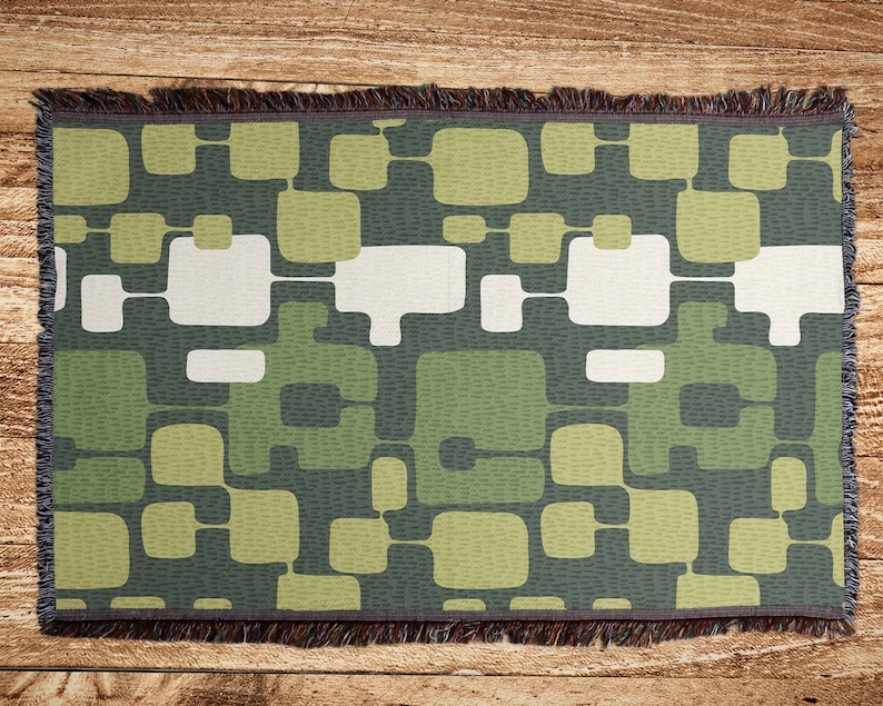 May include: A woven throw blanket with a geometric pattern of green, white, and gray squares and rectangles. The blanket has a fringe border.