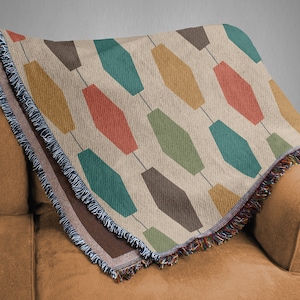 Pu&ograve; includere: Una coperta in tessuto tessuta con un motivo geometrico di esagoni allungati colorati su uno sfondo crema. La coperta ha un bordo a frangia.