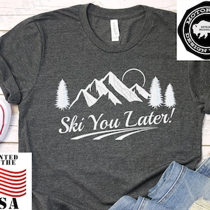 Op de afbeelding: Donkergrijs t-shirt met een witte berg- en boomafbeelding en de tekst "Ski You Later!". Het shirt heeft ook een "Printed in the USA" afbeelding.