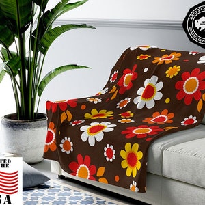 May include: A brown fleece throw blanket with a retro floral pattern featuring white, orange, and yellow flowers on a brown background. The blanket is draped over a white couch.