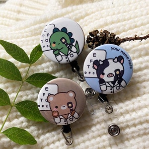 May include: Three round retractable badge holders with cartoon animal optometrists. The designs include a green dinosaur, a brown bear, and a black and white cow, each holding an eye exam tool. The text "your name here" is on one.