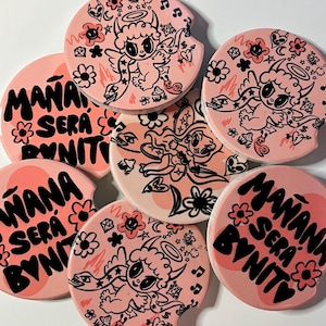 May include: A set of pink car coasters with a whimsical design. Each coaster features a cartoon angel with a halo, flowers, and the phrase "MAÑANA SERÁ BONITO" in black and pink lettering. The coasters are round.