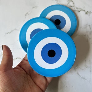 Könnte beinhalten: Drei runde, blaue Untersetzer mit einem Augendesign. Jeder Untersetzer hat einen hellblauen Hintergrund, einen weißen Ring, einen dunkelblauen Kreis und eine schwarze Mitte. Die Untersetzer sind auf einer weißen Marmoroberfläche angeordnet.