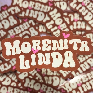 May include: Brown sticker with the words "Morenita Linda" in white, retro-style font. Small pink hearts accent the design. The sticker has a wavy border and is surrounded by similar stickers.