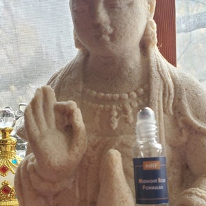 May include: A beige-colored statue of a seated figure with a serene expression, holding a small bottle. The statue has intricate details and is positioned near a window. The bottle has a blue label.