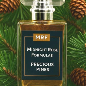 May include: A rectangular perfume bottle with a clear, faceted cap and a dark navy label. The label reads "MRF Midnight Rose Formulas Precious Pines." The bottle is filled with a golden liquid, set against a backdrop of pine needles and pine cones.